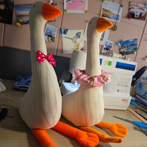 Two handmade sock geese with orange beaks and feet, one wearing a pink polka-dot bow tie and the other a pink plaid bow tie, displayed on a sewing table with a sewing machine in the background.
